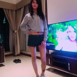 Woman posing indoors in a living room, wearing a gray cropped hoodie, dark shorts, and white sneakers, with a TV nearby.