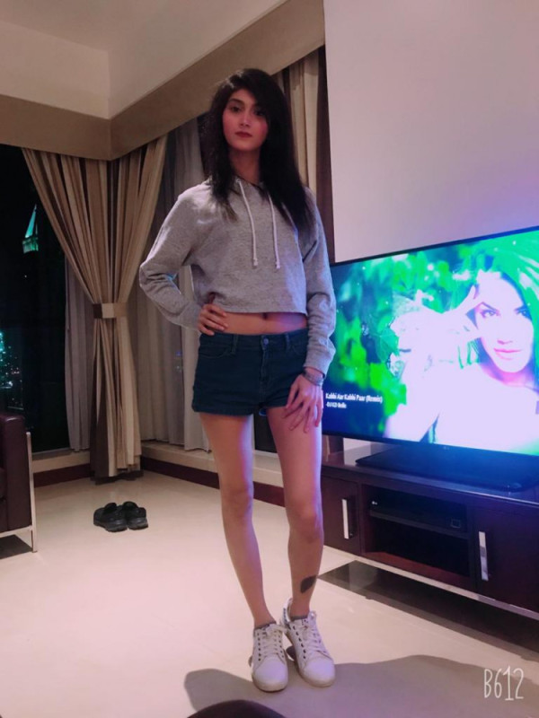 Woman posing indoors in a living room, wearing a gray cropped hoodie, dark shorts, and white sneakers, with a TV nearby.
